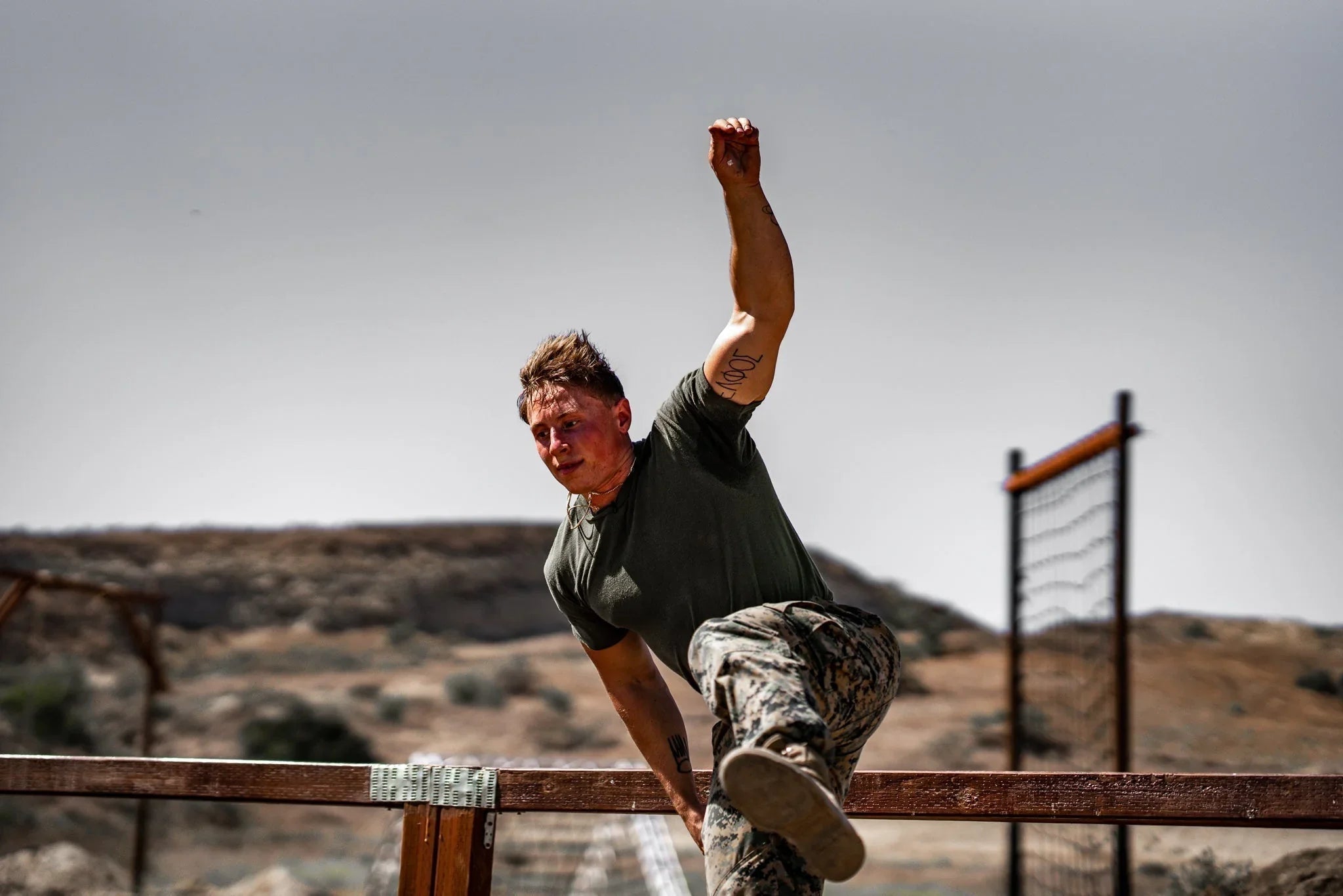 Military man in camouflage pants climbing over wooden battle bunker obstacle in arid outdoor training area