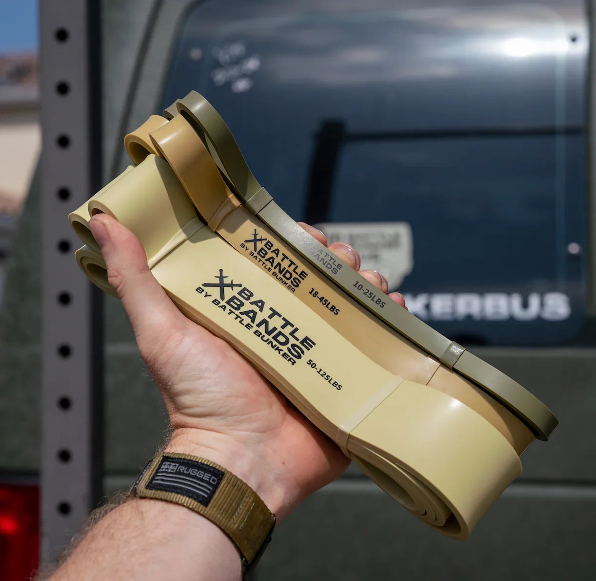 Hand holding three tan and olive Battle Bands resistance bands by Battle Bunker with vehicle in background