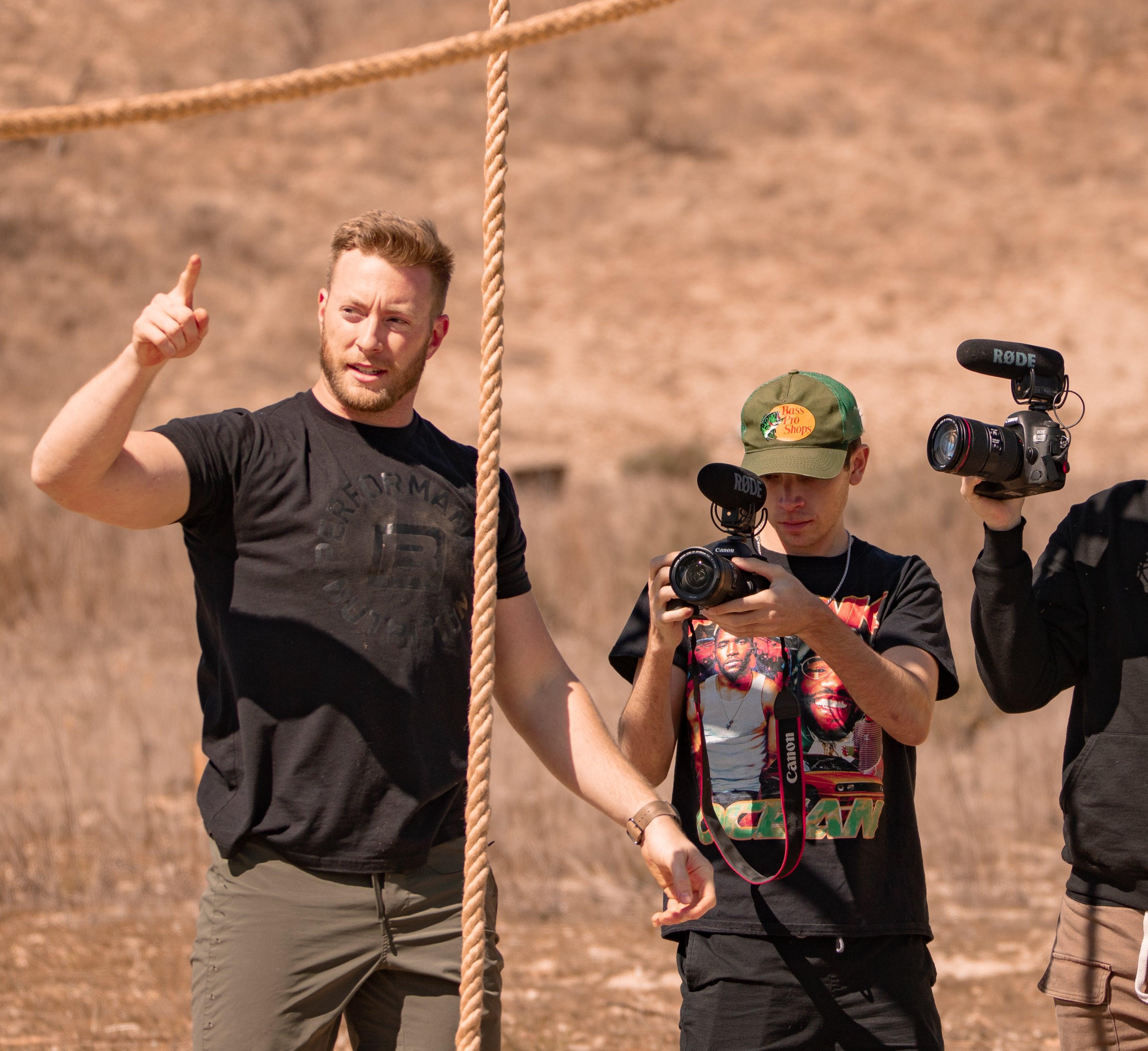 Man gesturing with finger while two cameramen film outdoors at battle bunker site