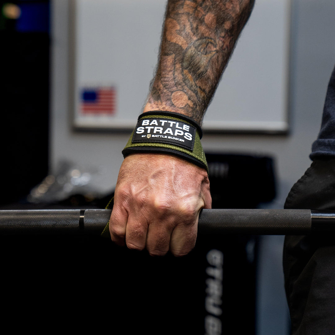 Close-up of a tattooed hand gripping a barbell with green Battle Straps wrist wrap by Battle Bunker