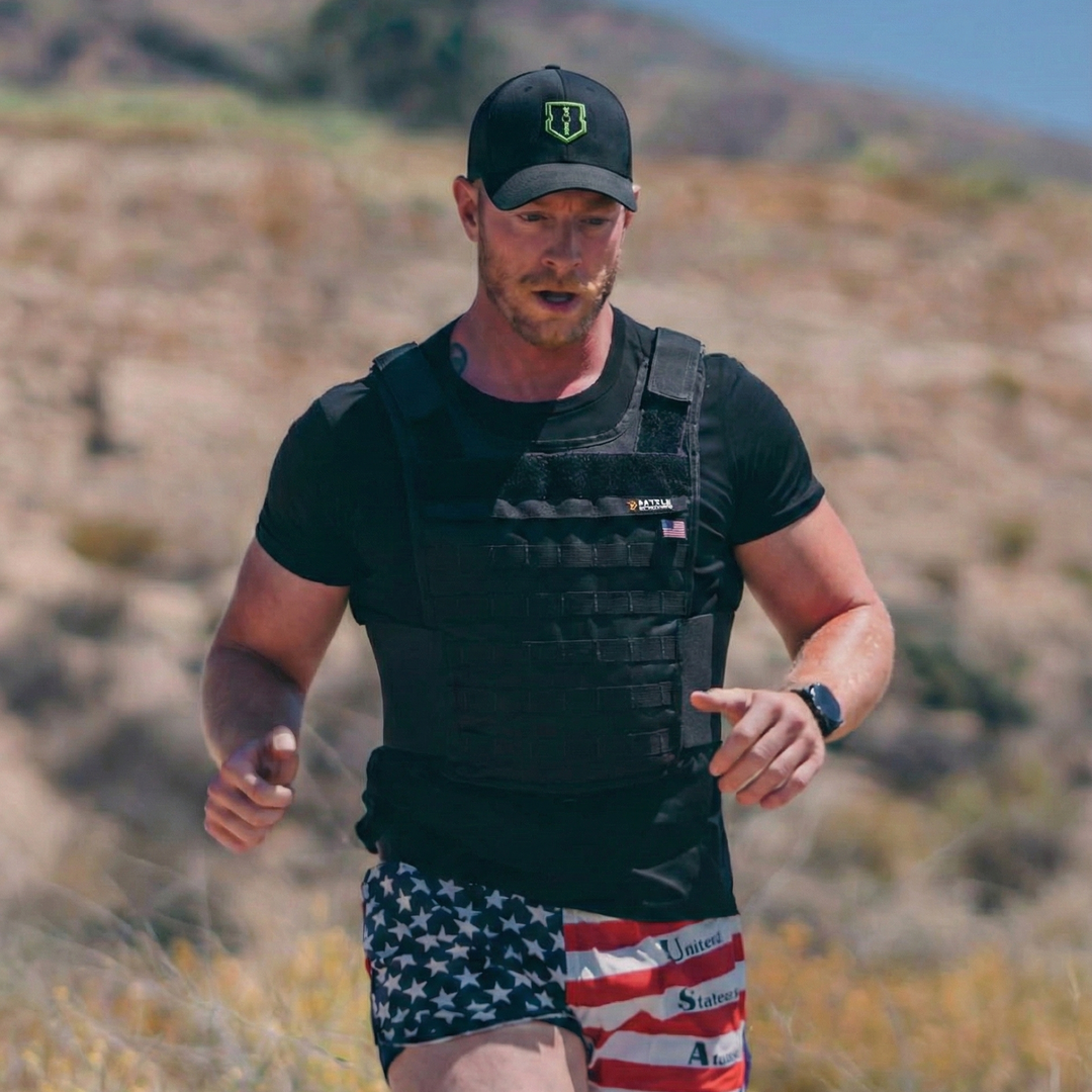 Man running outdoors wearing a black tactical vest and American flag shorts.