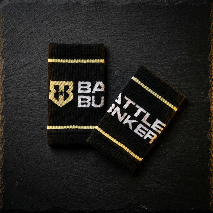 Black Battle Bunker wristbands with gold stripes and logo on dark textured background